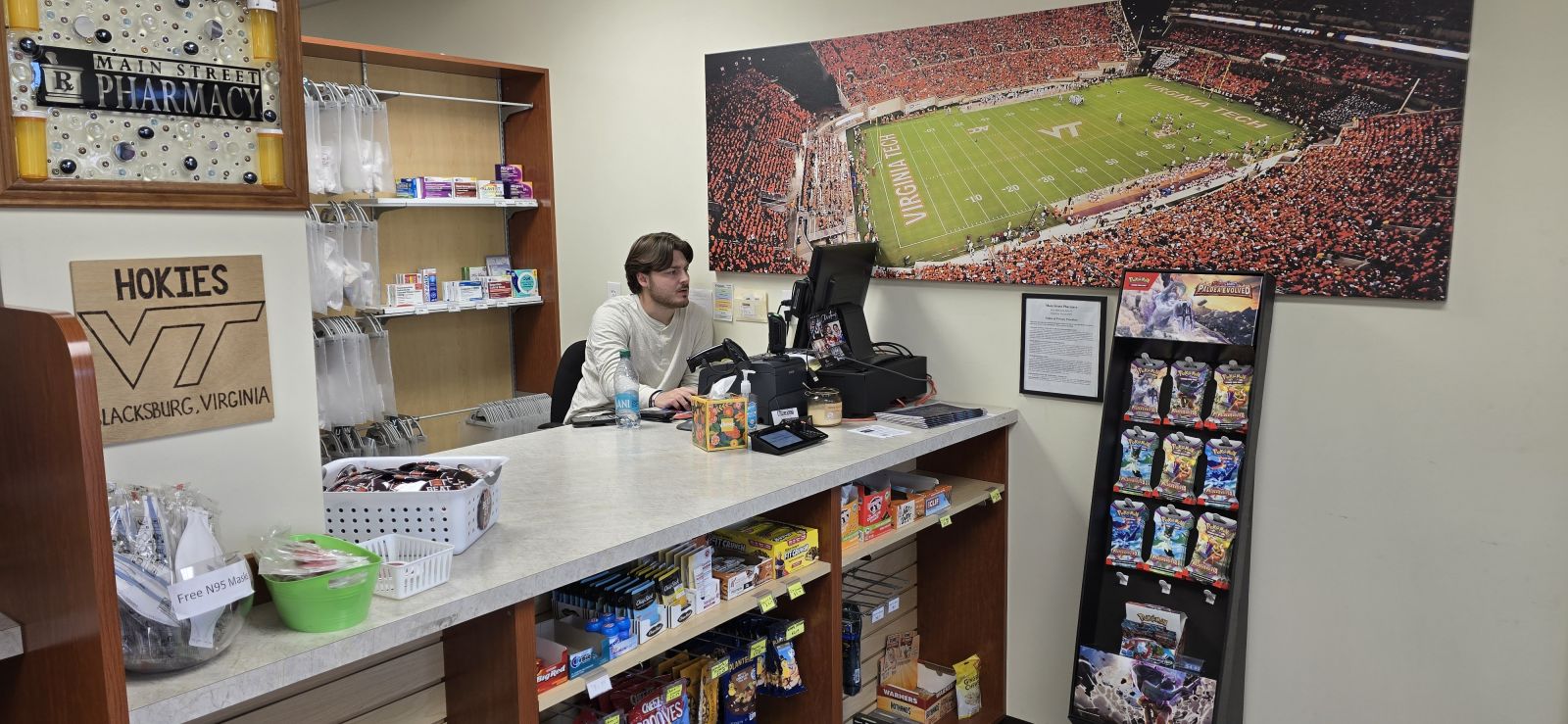 A man works at a pharmacy counter with shelves of medicine and snacks, and a large football stadium photo on the wall.