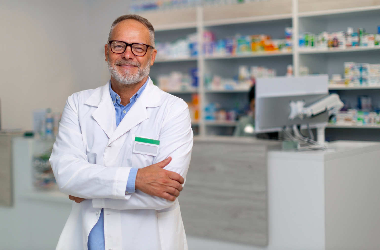 2150600097 Smiling pharmacist in white coat standing with arms crossed in a pharmacy.