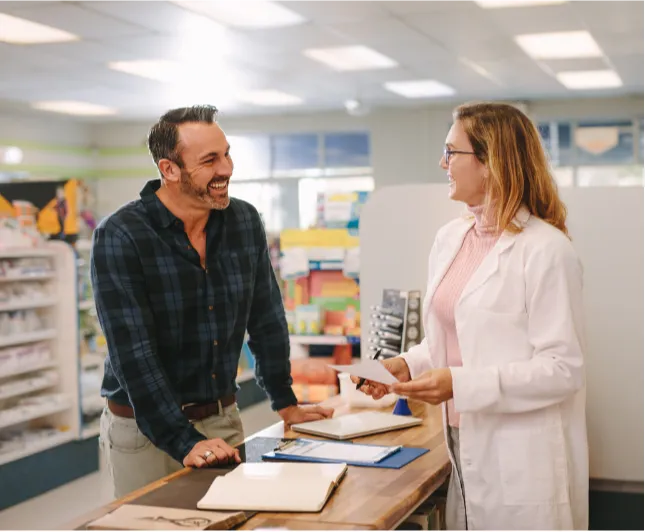 25-50-1 A smiling man talks with a female pharmacist at a pharmacy counter, holding a prescription paper.