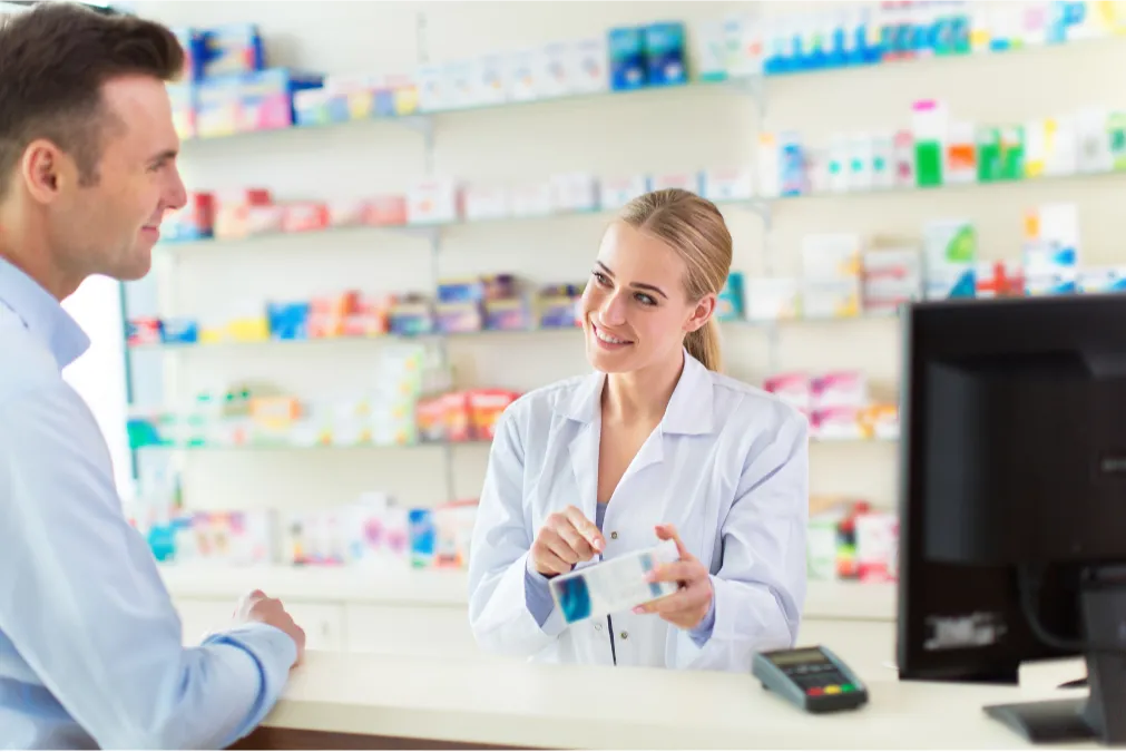 25-50-2 A smiling pharmacist helps a customer at a pharmacy counter, holding a box of medication.