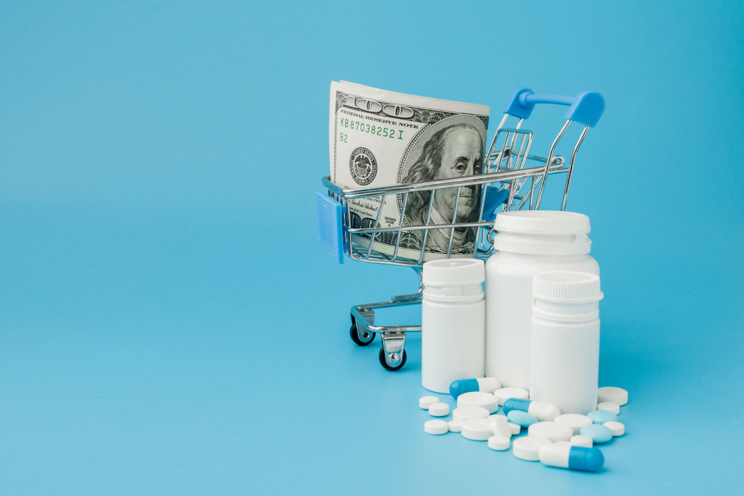 Mini shopping cart with cash, pill bottles, and scattered pills on a blue background.