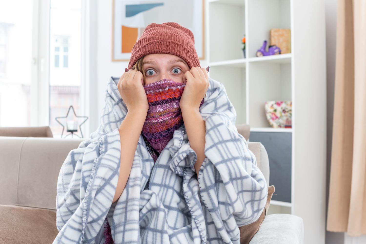 Person bundled in a blanket and hat, looking surprised with scarf covering mouth, sitting on a couch indoors.