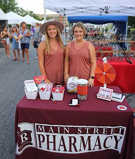 about-community Two women stand behind a Main Street Pharmacy table with prizes at an outdoor community market event.