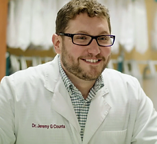 A smiling man in glasses and a white lab coat stands in a medical or laboratory setting.